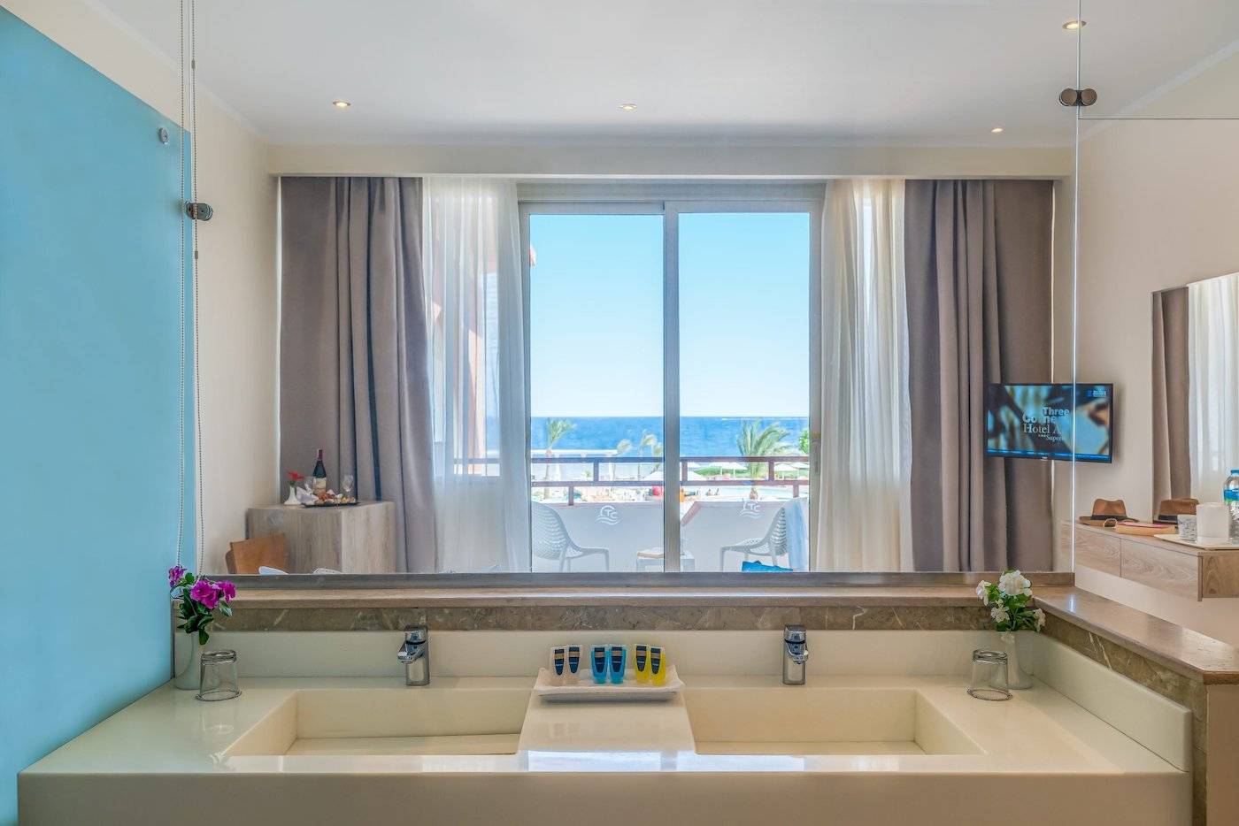 Badezimmer-Ansicht im Three Corners Equinox Beach Resort mit großem Fenster, das auf den Ozean und eine Terrasse mit Stühlen hinausblickt.