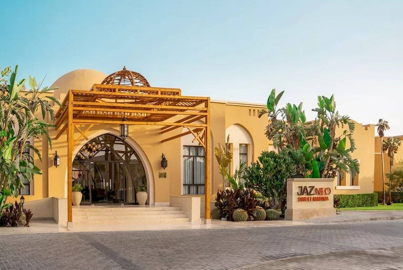 Außenansicht des Jaz Neo Dar El Madina Hotels mit gelben Wänden, architektonischem Eingang und grünen Pflanzen davor.