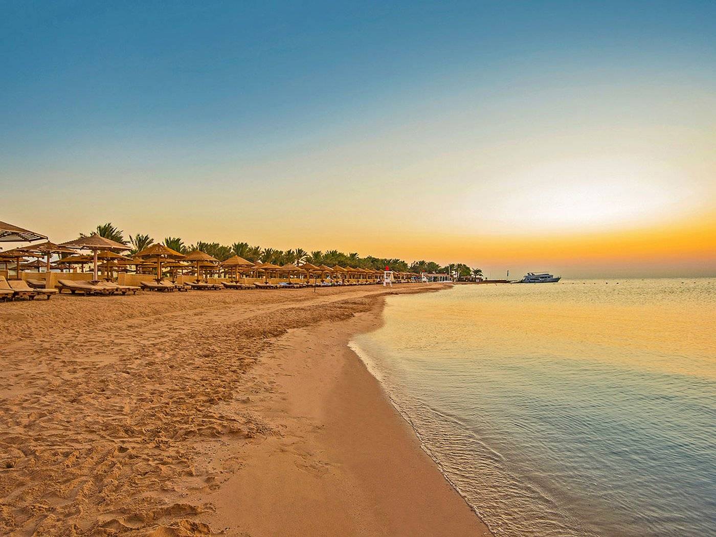 Sonnenuntergang am Strand des SUNRISE Royal Makadi Resort mit leeren Sonnenliegen und einem Boot im Wasser.