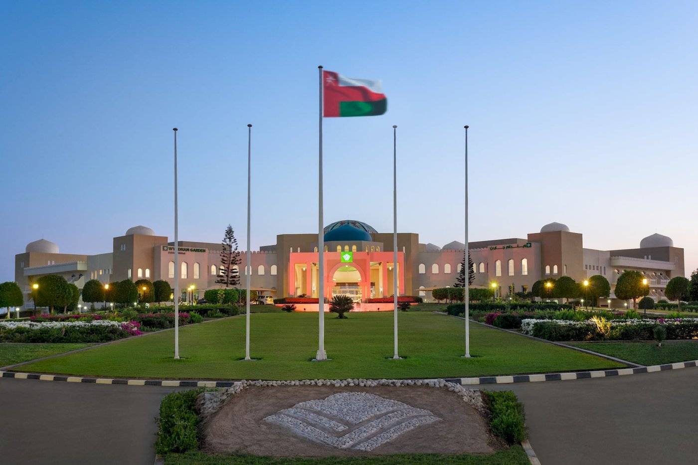 Aussenansicht des Wyndham Garden Salalah Mirbat Hotels mit Flaggen und beleuchteter Fassade