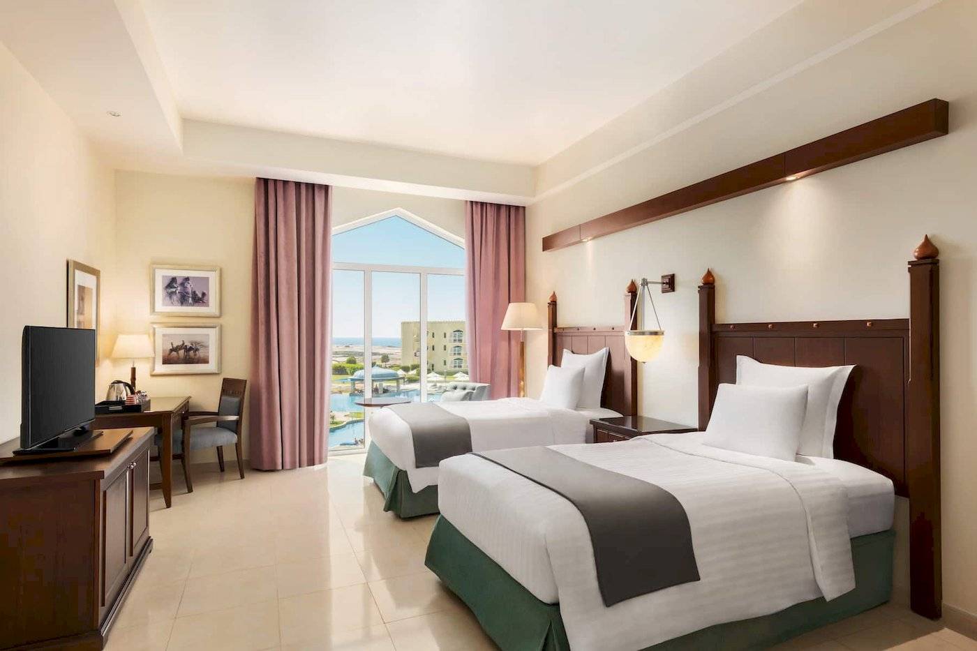 Doppelzimmer mit Meerblick im Wyndham Garden Salalah Mirbat Hotel, mit zwei Betten, Fernseher und Tisch, sowie ein großer Fenster mit Poolsicht.