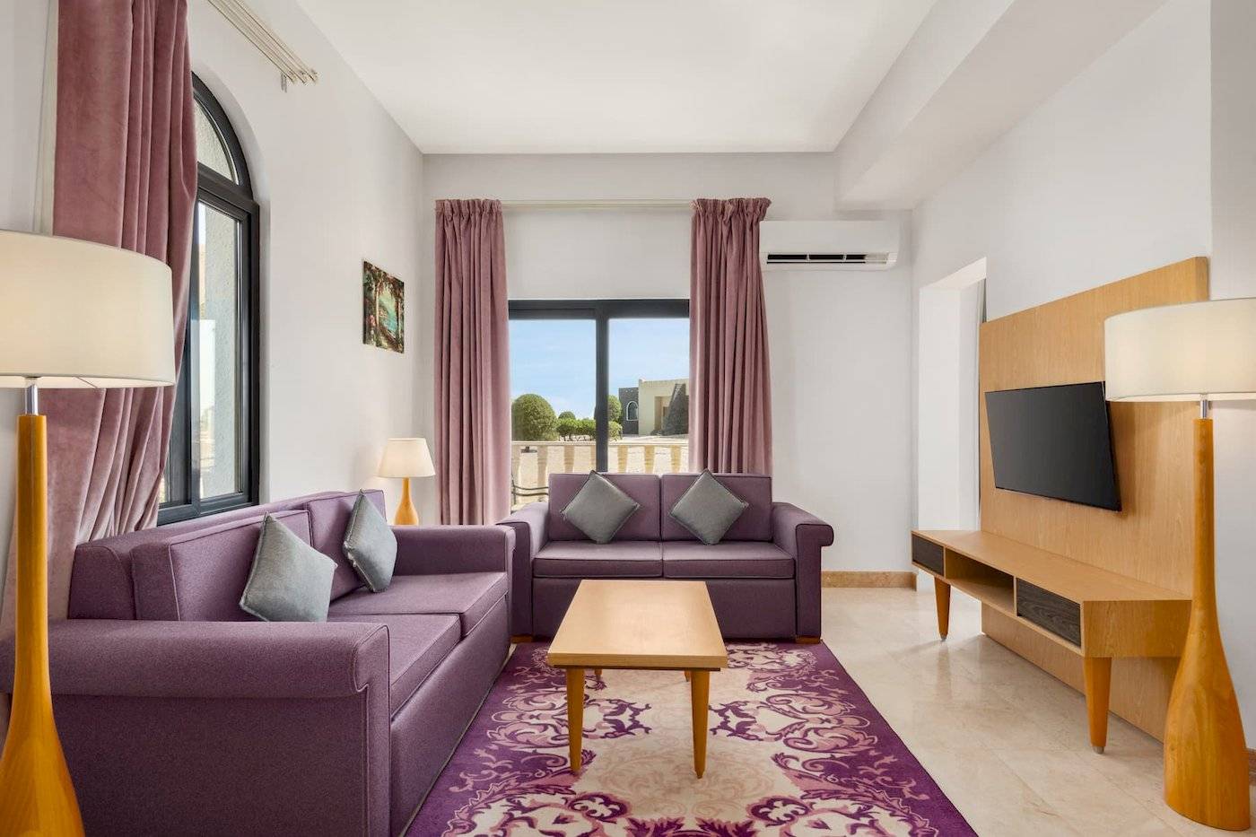 Wohnzimmer im Wyndham Garden Salalah Mirbat mit zwei lila Sofas, einem Tisch, Lampen und einem Fernseher.