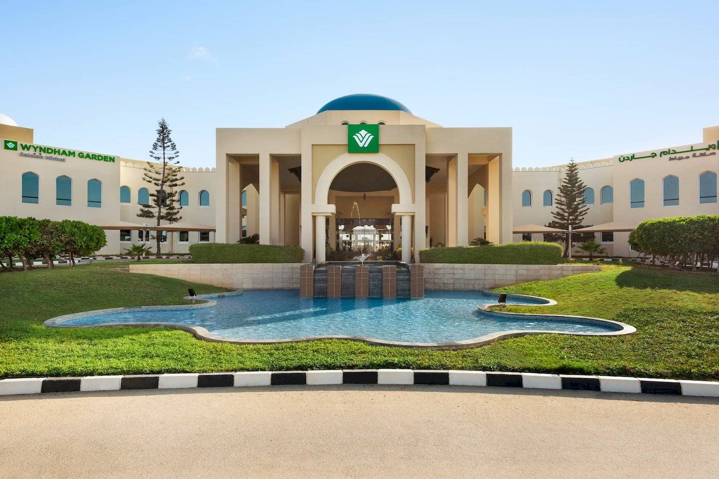 Außenansicht des Wyndham Garden Salalah Mirbat Hotels mit Brunnen und Rasenfläche.