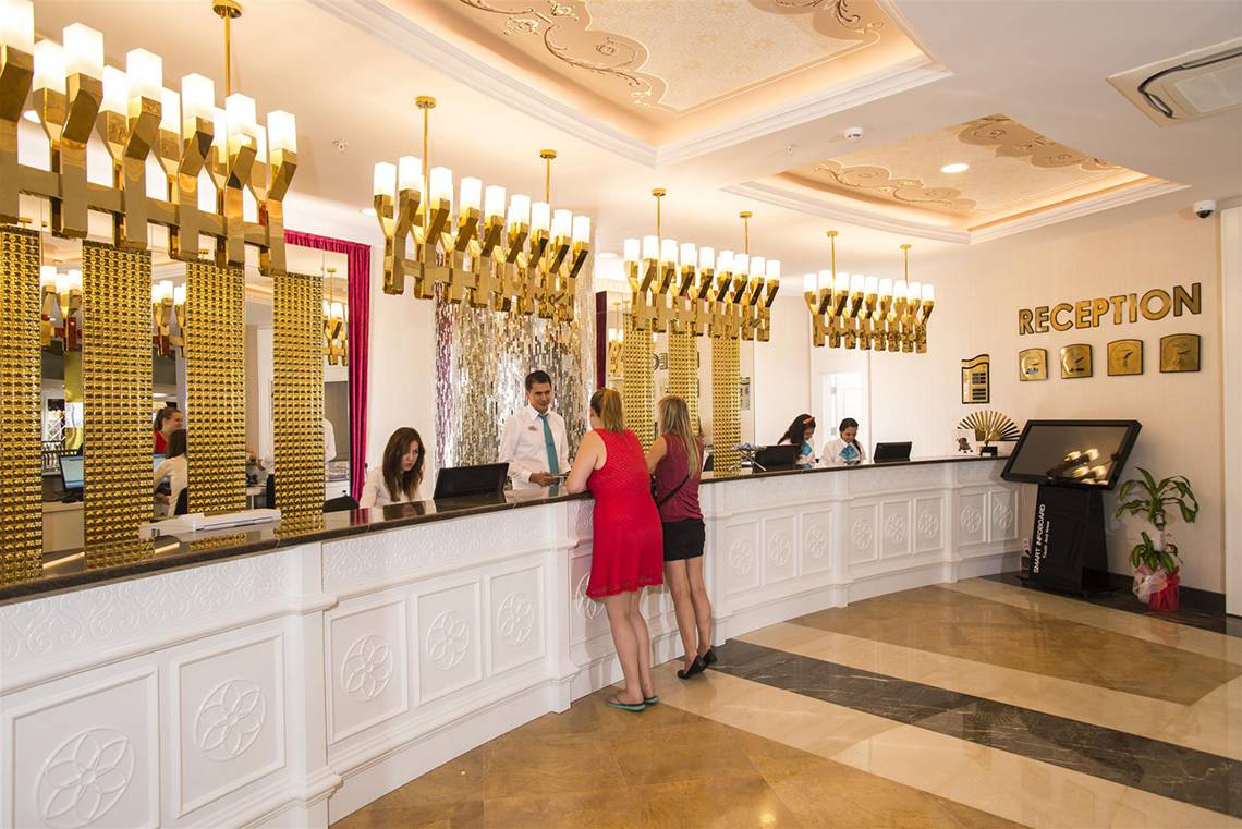 Rezeptionsbereich des Dream World Aqua Hotels mit goldenen Kronleuchtern und einer Rezeptionistin, die zwei Gäste bedient.