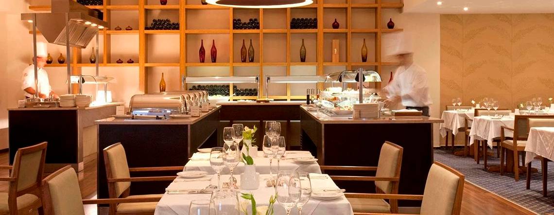 Innenbereich des Alion Beach Hotel Restaurants mit gedeckten Tischen, Geschirr und Köchen hinter einer Theke.