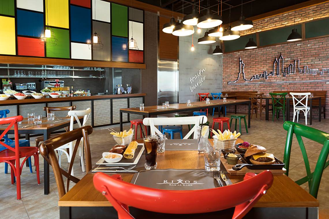 Innenansicht des Rixos Bab Al Bahr Restaurants mit bunten Stühlen und Tischen, serviertem Essen und einer Wand mit Toast Burger-Grafik.