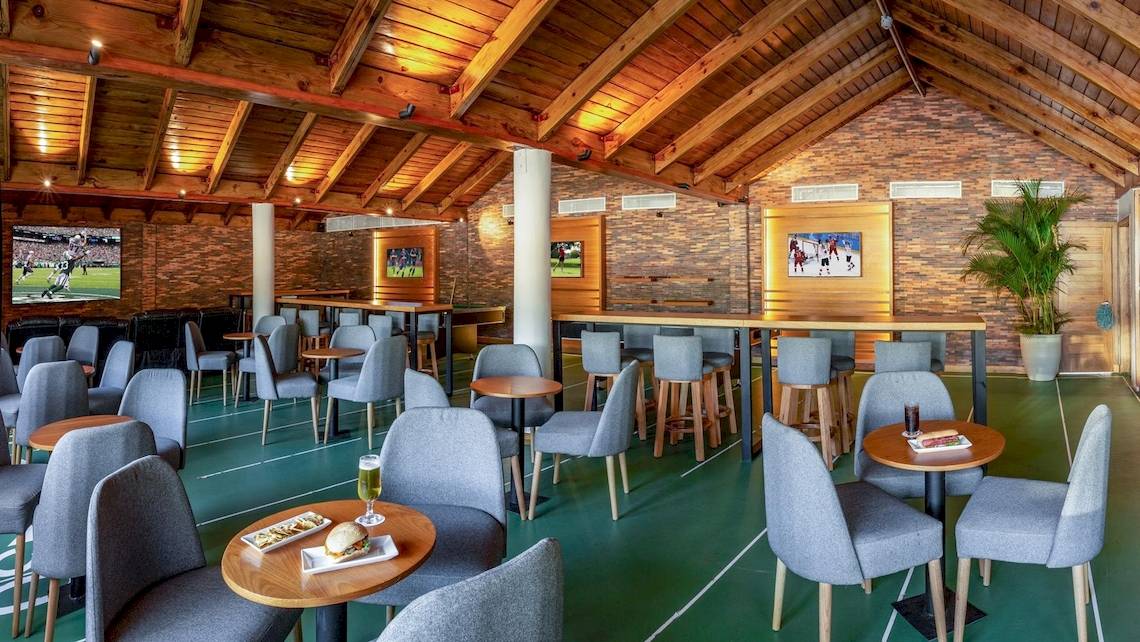 Innenbereich des Grand Sirenis Punta Cana Resort mit mehreren Tischen und Stühlen, Fernsehern an der Wand und einer grünen Spielfläche.