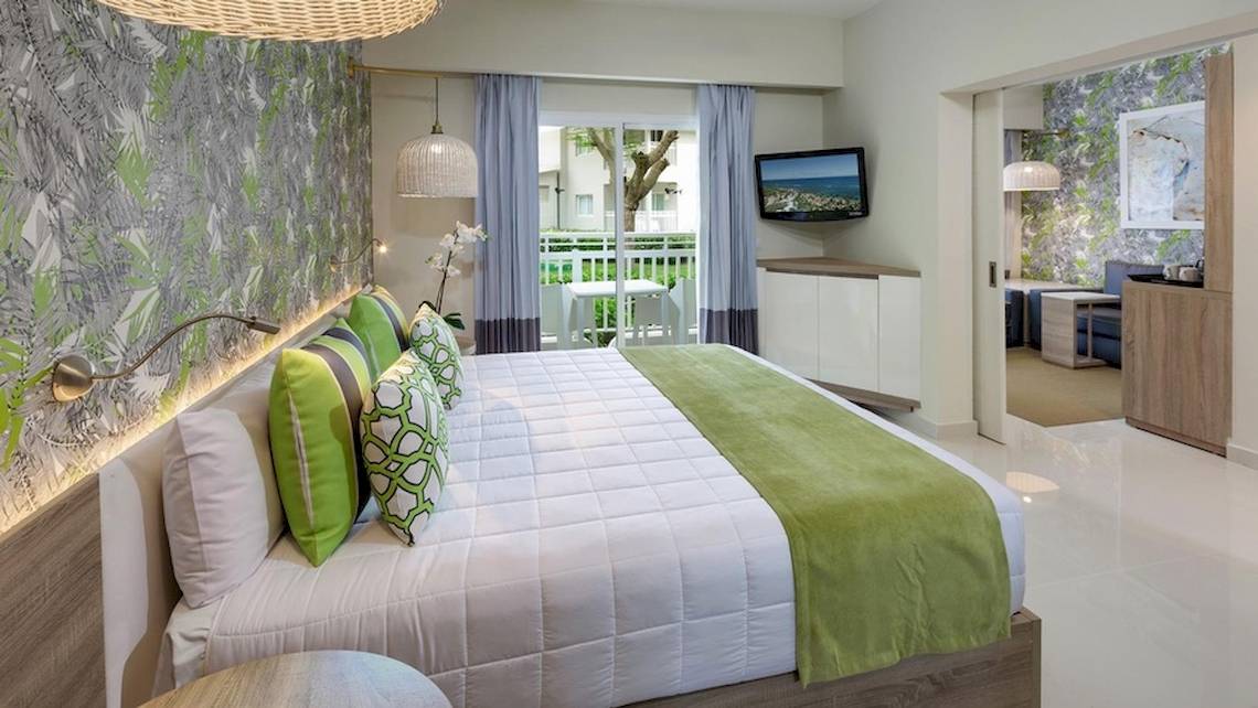 Innenansicht eines Schlafzimmers im Grand Sirenis Punta Cana Resort mit einem großen Bett, grünen Kissen und einem Fernseher an der Wand.