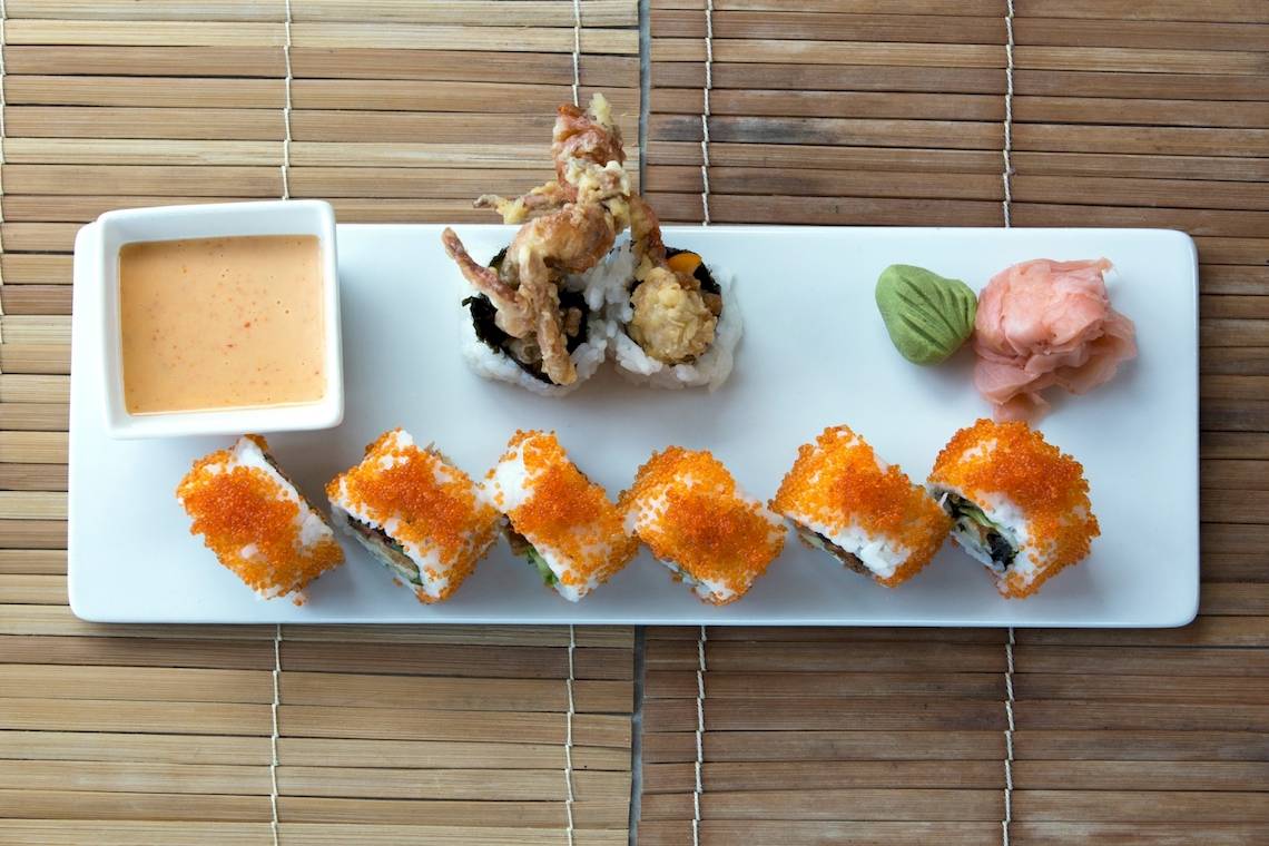 Ein weißes Tablett mit verschiedenen Sushi-Rollen und einer kleinen Schüssel Sauce auf einer Bambusmatte.