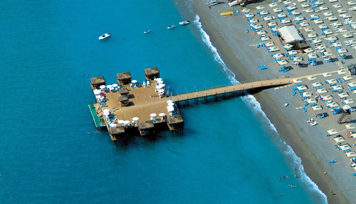 Luftaufnahme des Club Nena Hotels mit Außenbereich am Strand, vielen Sonnenschirmen und einem Steg zum Wasser.