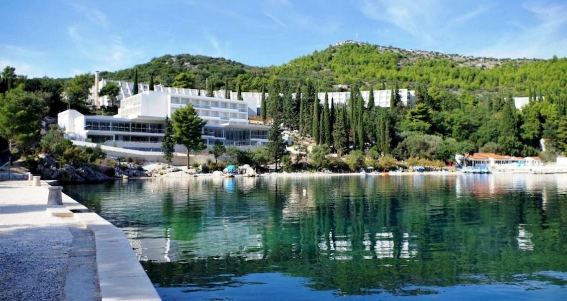 Außenansicht des Osmine Slano Hotels am Wasser mit grünen Bäumen und Gebäuden im Hintergrund.