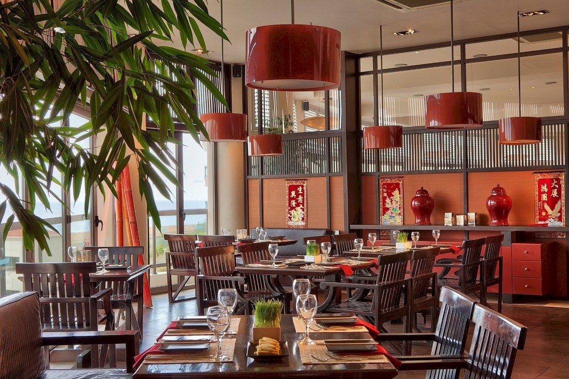 Innenansicht des Restaurants im Ikos Oceania Hotel mit runden roten Lampen und Holzmöbeln.