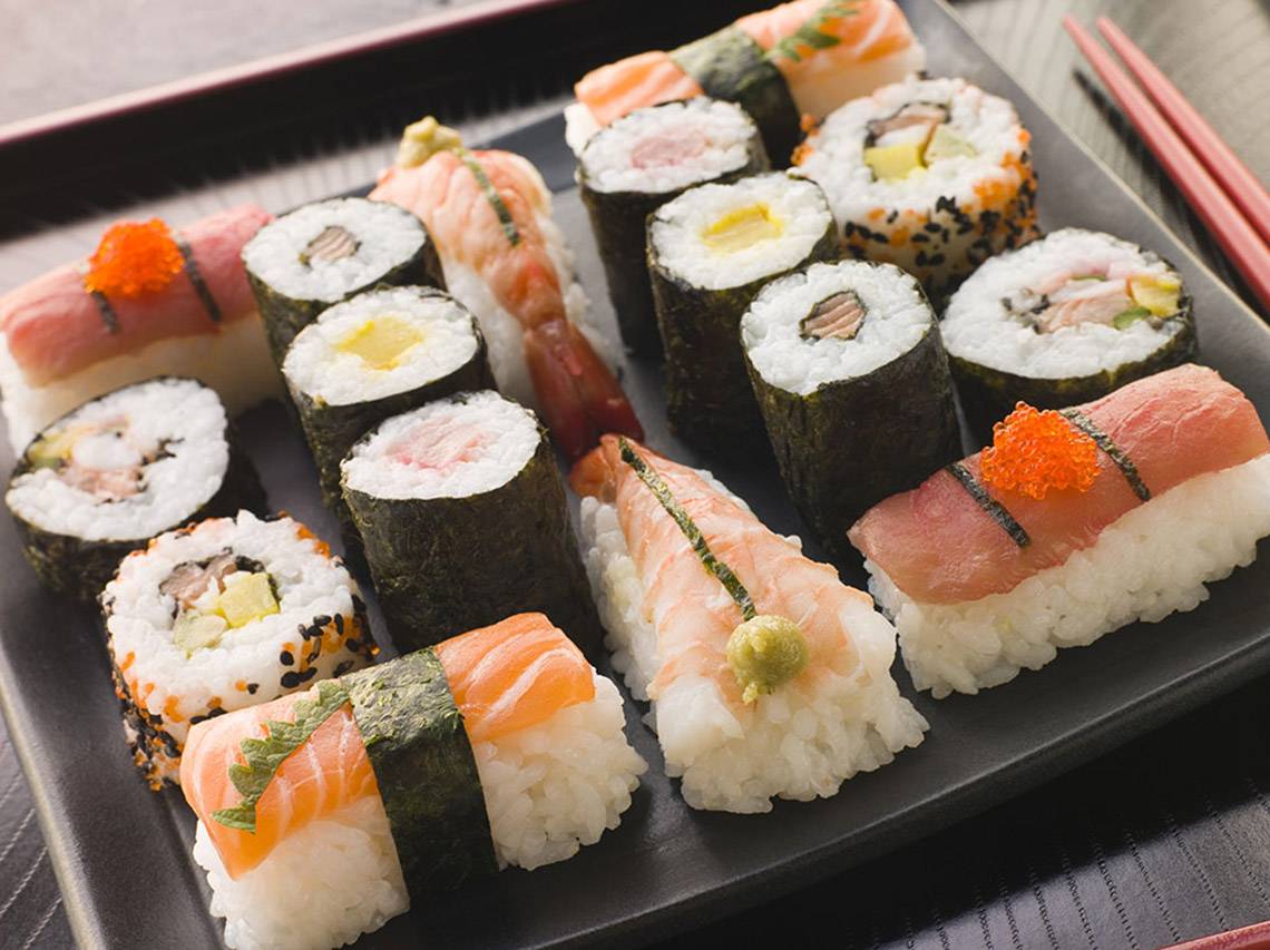 Eine Auswahl verschiedener Sushi-Rollen auf einem schwarzen Teller, mit Stäbchen daneben.
