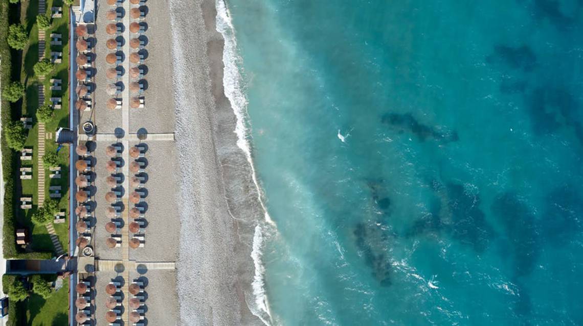 Luftaufnahme des Rhodes Bay Hotel & Spa Strandes mit vielen Sonnenschirmen und klarem blauem Wasser.