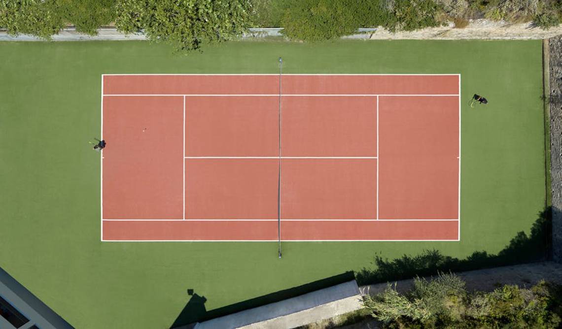 Luftaufnahme des Tennisplatzes im Rhodes Bay Hotel & Spa mit zwei Spielern auf grünem und rotem Boden.