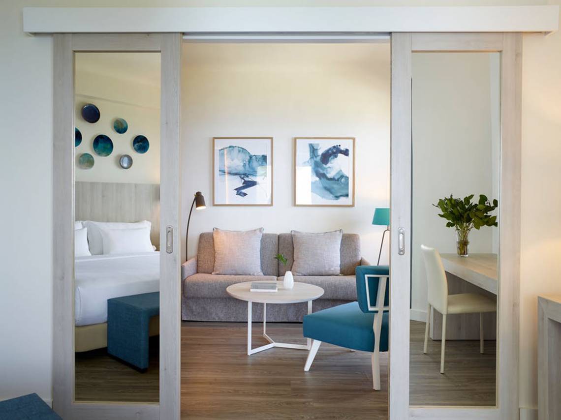 Inneneinrichtung von einem Doppelzimmer im Rhodes Bay Hotel & Spa mit einem Bett, Sofa, Tisch, Stuhl und Lampe.
