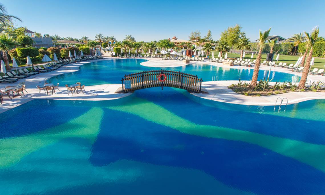 Pool des Sherwood Exclusive Lara Hotels mit Sonnenliegen, Schirmen und einer Brücke.