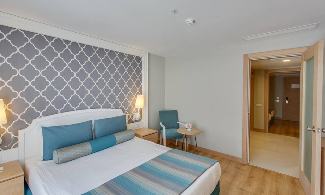 Innenansicht eines Schlafzimmers im Sherwood Exclusive Lara Hotel mit Bett, Nachttischen und offenem Tür.