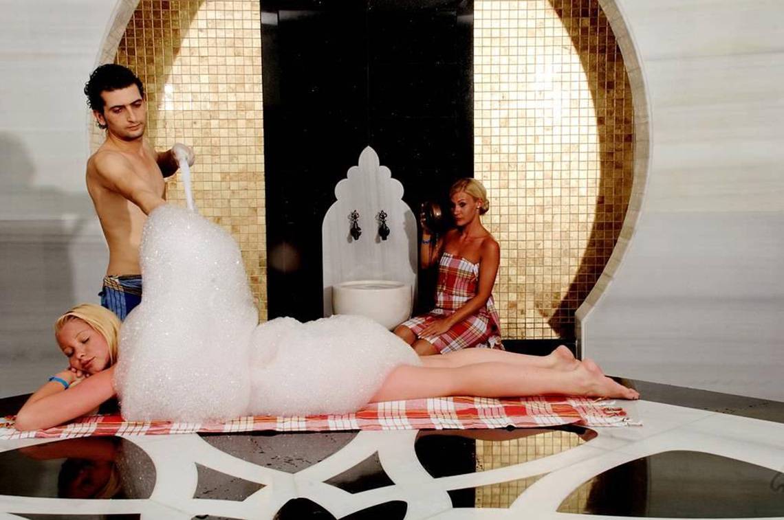 Mann und Frauen im Hammam mit Seifenblasen, ein Mann gießt Seifenblasen auf eine schlafende Frau