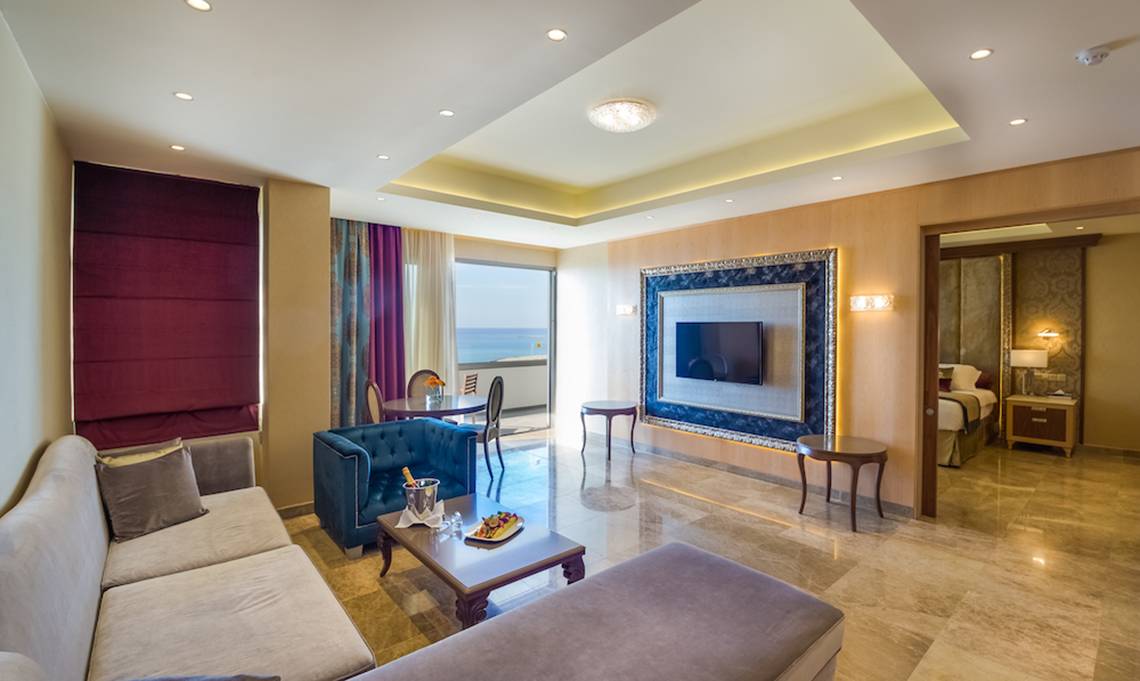 Innenansicht eines Zimmers im Adams Beach Hotel mit Wohnzimmer, Essbereich, Fernseher und Blick auf das Meer.