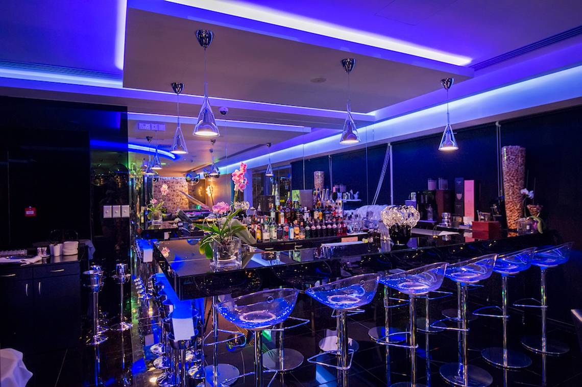 Die Bar des Adams Beach Hotels mit moderner Einrichtung, blauer Beleuchtung und transparenten Stühlen.