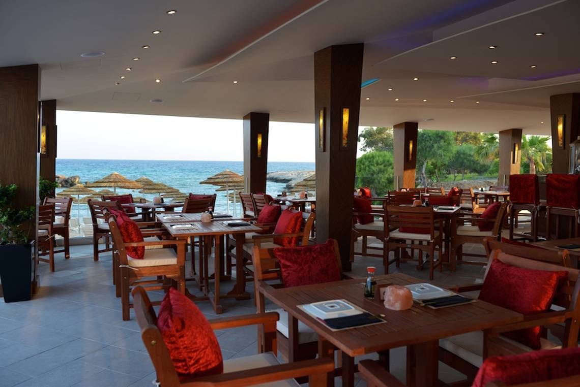 Innenansicht des Restaurants im Adams Beach Hotel mit Blick auf das Meer und Sonnenschirme.