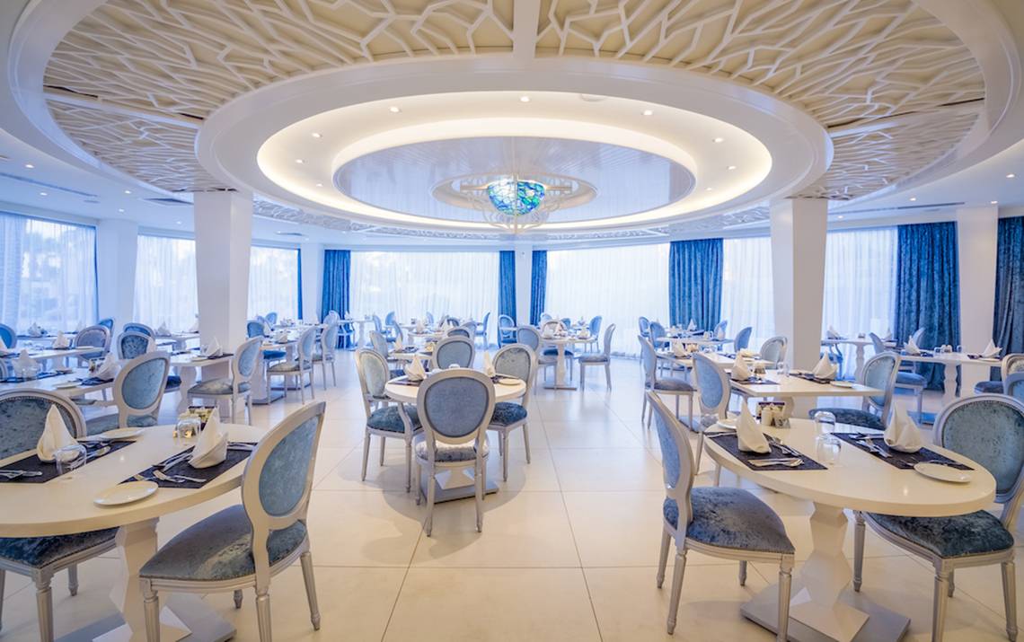 Innenbereich des Adams Beach Hotels, Restaurant mit runden Tischen und Stühlen, zentrale Beleuchtung und blaue Vorhänge.