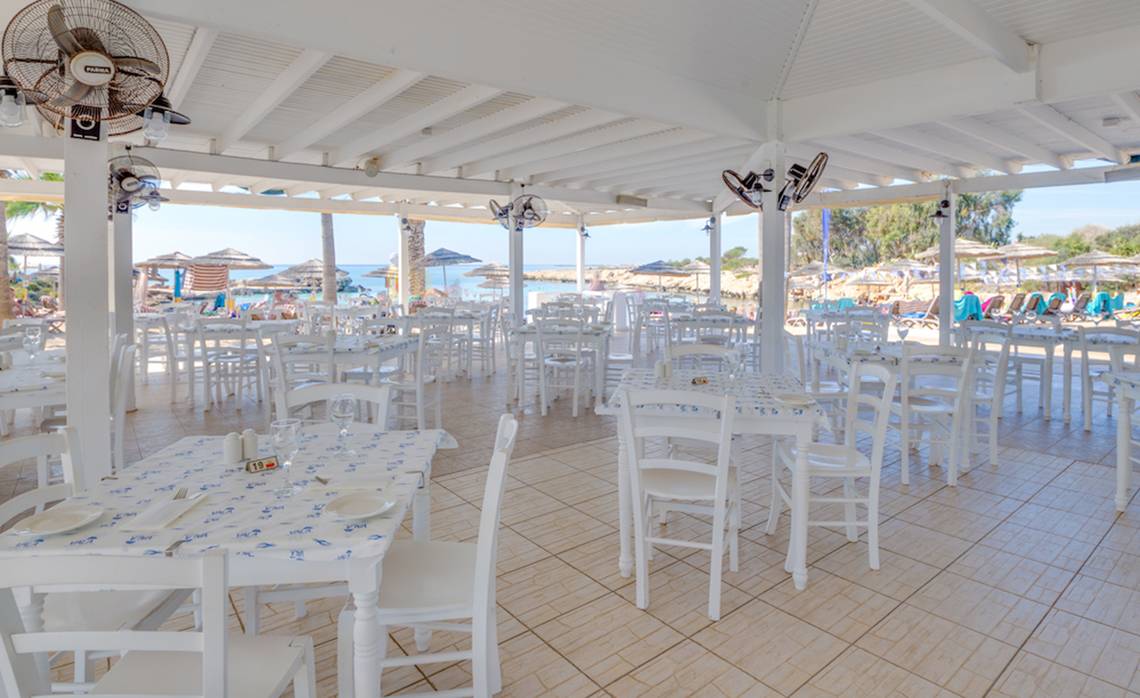 Innenbereich des Adams Beach Hotel Restaurants mit weißen Tischen und Stühlen und Blick auf den Strand.