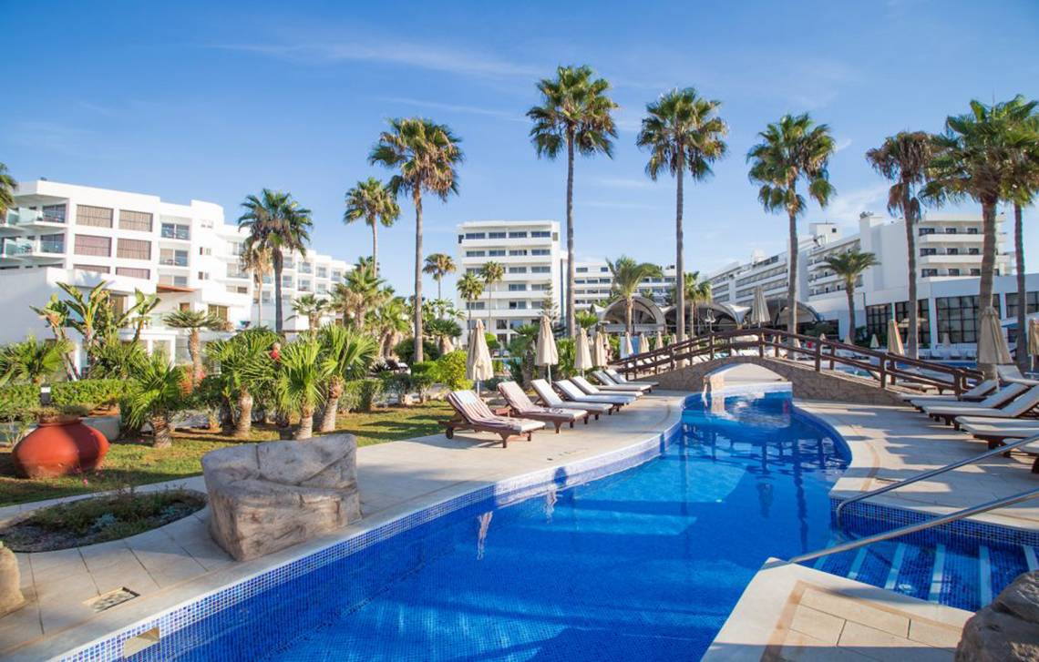 Außenpoolbereich des Adams Beach Hotels mit Palmen, Sonnenliegen und weißen Gebäuden im Hintergrund.