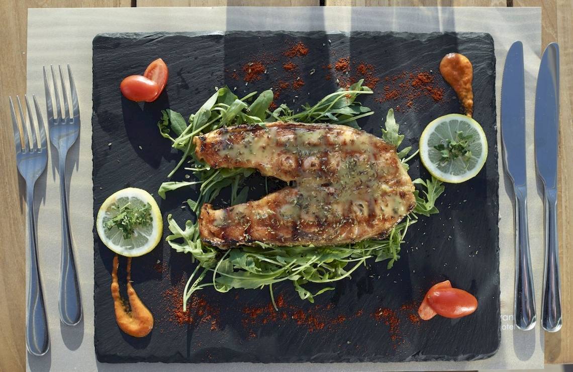 Gegrillter Fisch auf schwarzem Teller mit Rucola und Zitronenscheiben.