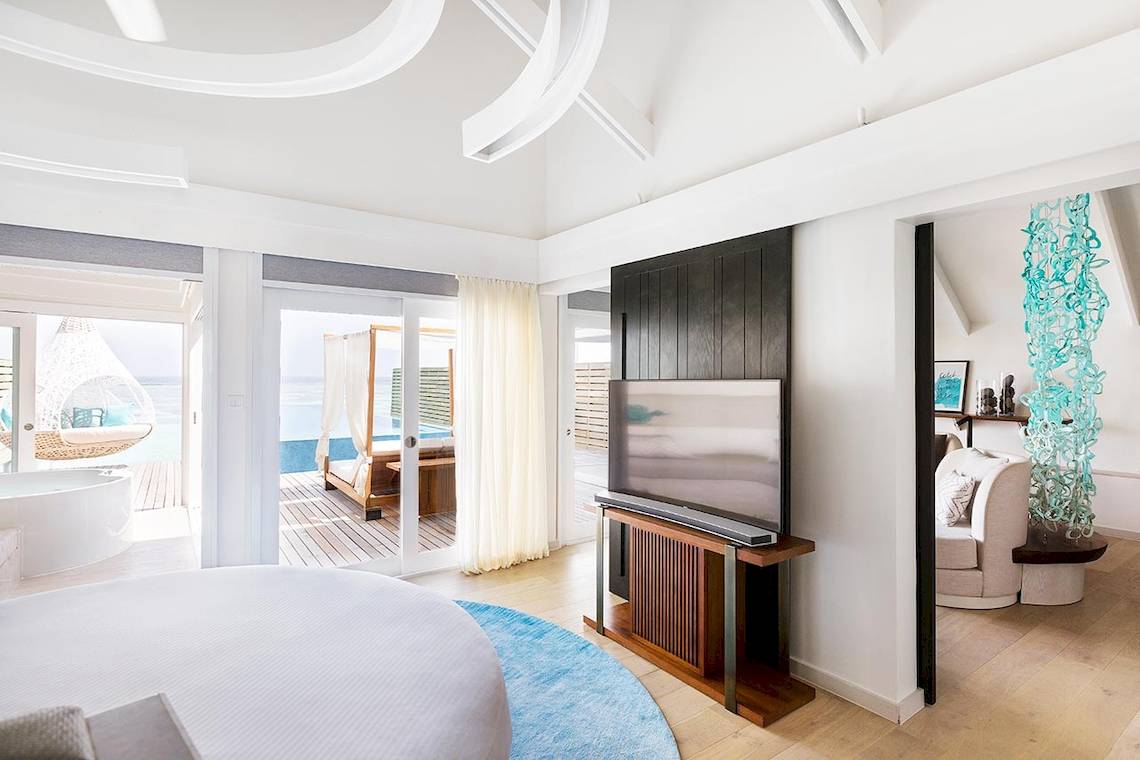 Innenansicht einer Suite im LUX South Ari Atoll Hotel mit großem Bett, Fernseher und Glastüren zum Balkon.