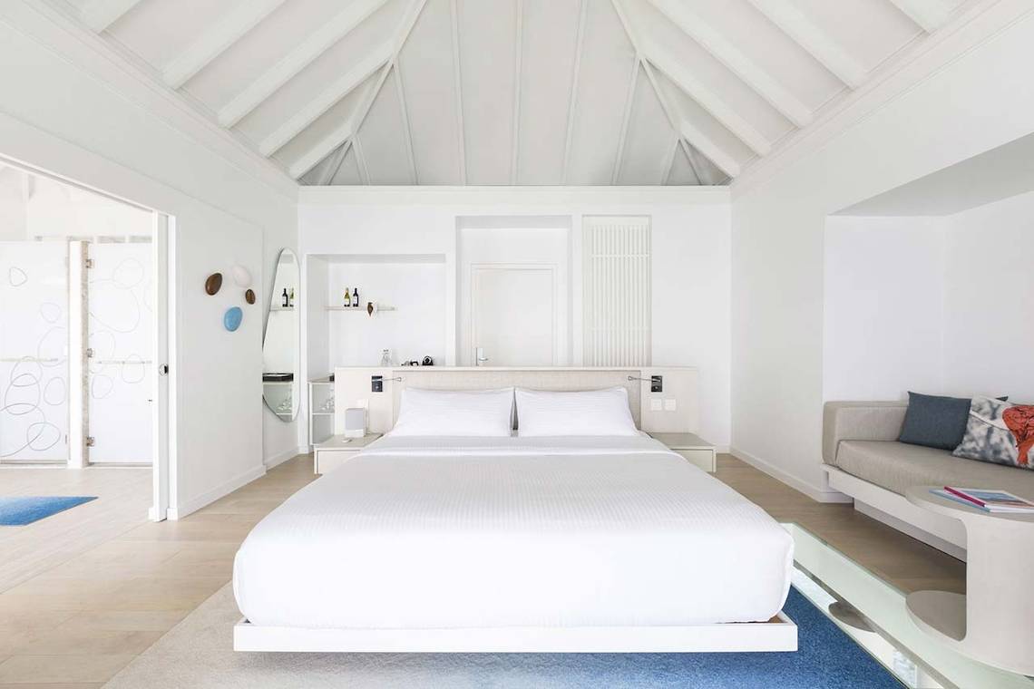 Innenansicht eines Schlafzimmers im LUX South Ari Atoll Hotel mit weißen Wänden und Möbeln, einem großen Bett und einer Couch.