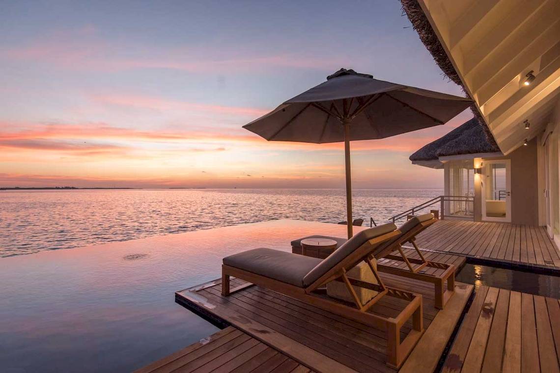 Terrasse des LUX South Ari Atoll Hotels mit zwei Liegen und einem Sonnenschirm am Meer bei Sonnenuntergang.