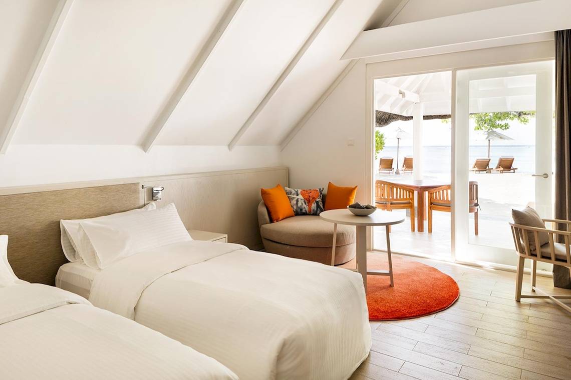 Innenansicht eines Doppelzimmers im LUX South Ari Atoll Hotel mit zwei Betten, einem kleinen Tisch und einer Aussicht auf den Strand.