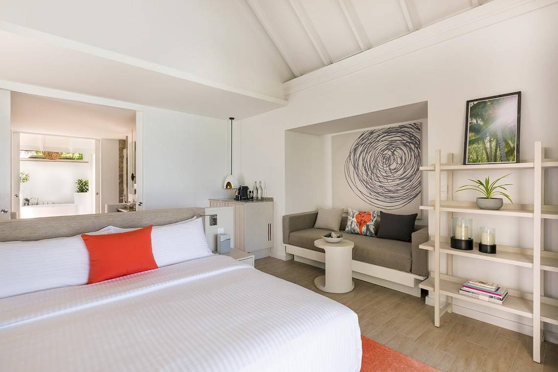 Innenansicht des Schlafzimmers im LUX South Ari Atoll Hotel mit Bett, Sofa, Nachttisch und Regal.