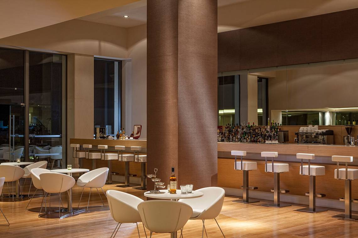 Innenansicht der Bar im Avra Imperial Hotel mit Holzboden, weißen Stühlen und einer Theke mit vielen Flaschen.