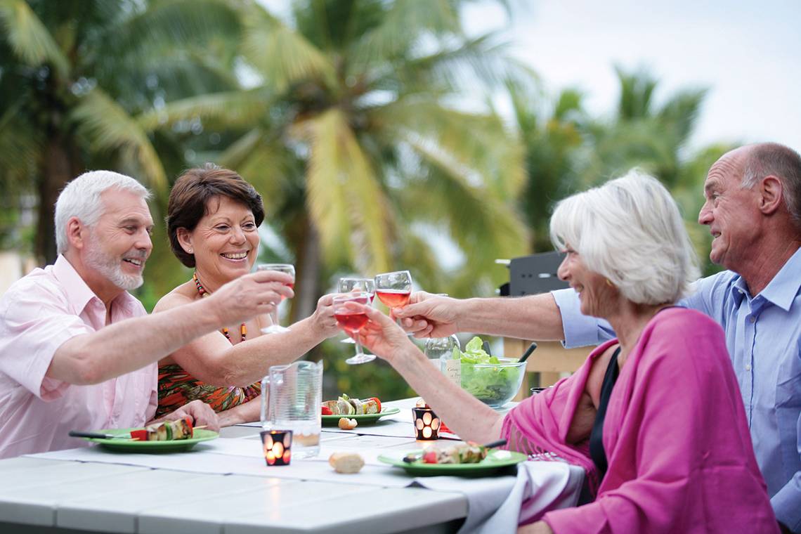 Gruppe älterer Menschen, die bei einer Feier draußen auf der Terrasse sitzen und Getränke anstoßen.
