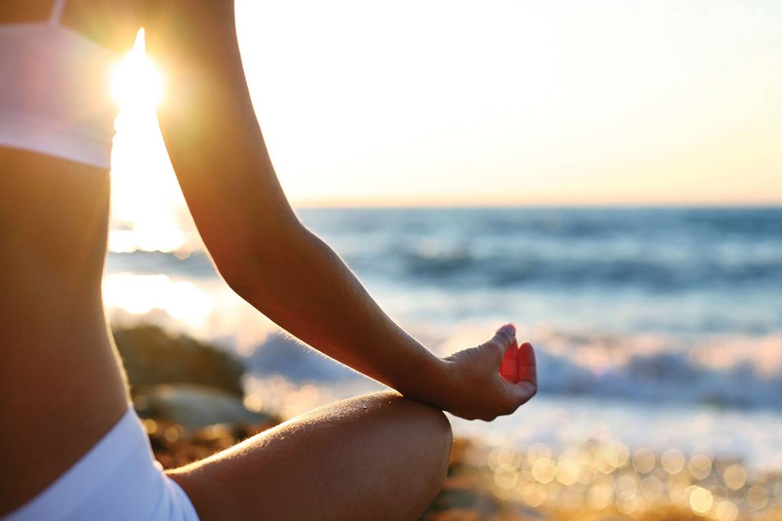 Frau in Meditation am Strand bei Sonnenuntergang mit dem Ozean im Hintergrund.