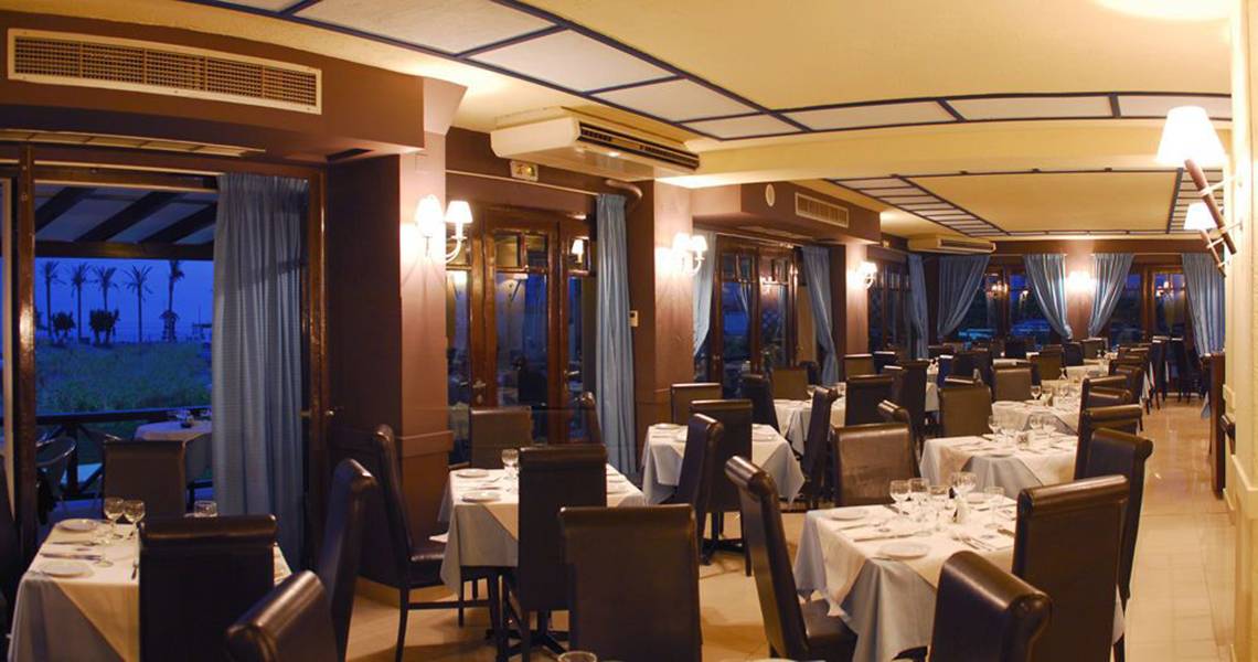 Innenansicht des Restaurants im Hersonissos Palace Hotel mit gedeckten Tischen und Stühlen, blauen Vorhängen und Lampen.