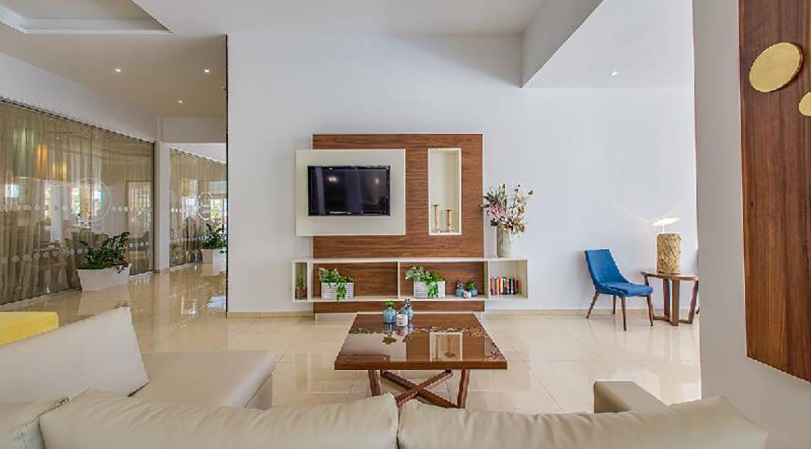 Modernes Wohnzimmer im Stamatia Hotel mit weißem Sofa, Glastisch und Fernseher an der Wand.