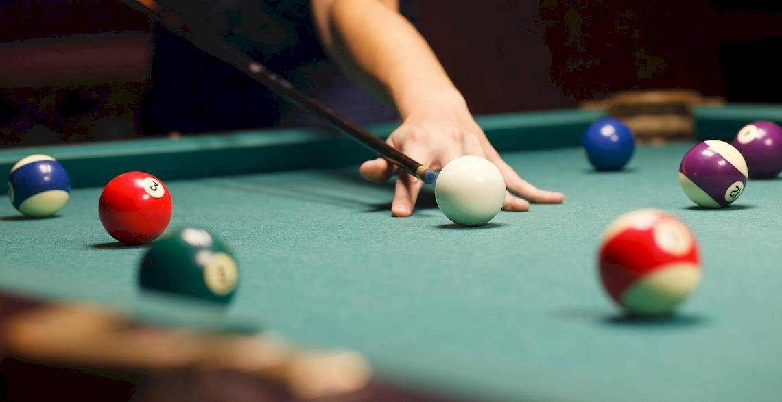 Eine Hand hält einen Poolstock und zielt auf eine weiße Kugel auf einem Pooltisch mit verschiedenen farbigen Kugeln.