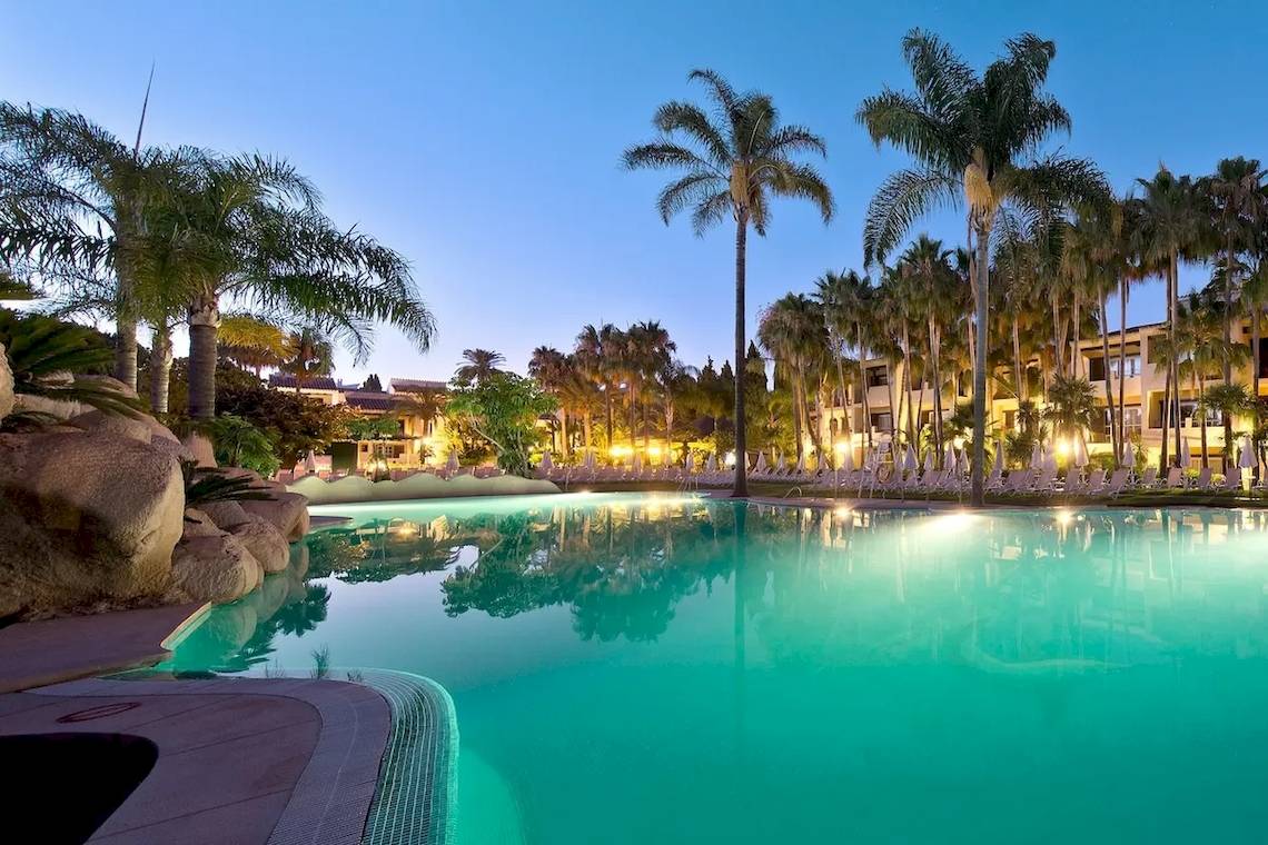 Abendansicht des Poolbereichs des Bluebay Banus Hotels mit leuchtenden Palmen und gesäumten Sonnenliegen.