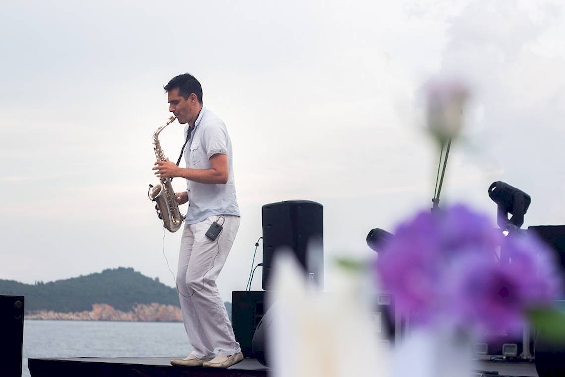 Mann spielt Saxophon auf Bühne mit Bokeh von Blumen und Lautsprechern im Hintergrund.