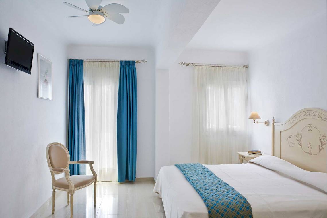 Schlafzimmer im Kamari Hotel mit weißem Bett, blauen Vorhängen und Wand-TV.
