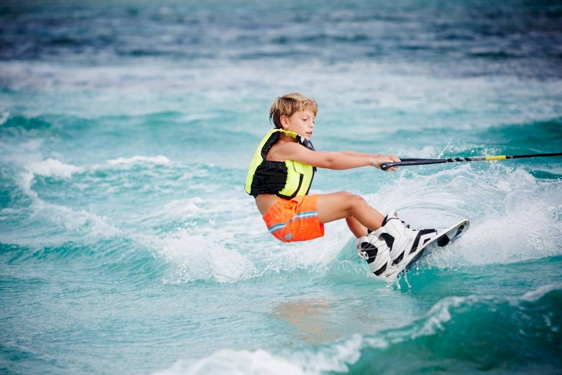 Junge im Ozean segelt auf Wellen, trägt orange Shorts und gelben Sicherheitsgurt.