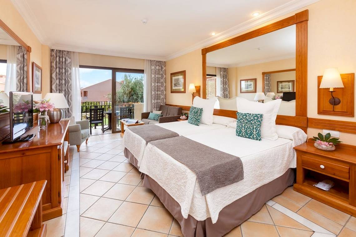 Doppelzimmer mit Balkon im GF Gran Costa Adeje Hotel, mit zwei Betten, Fernseher, Tisch und Stühlen auf dem Balkon.