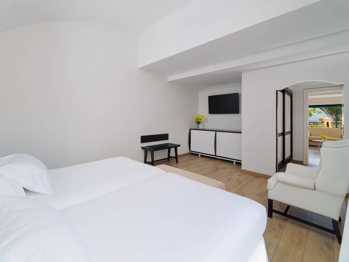 Schlafzimmer im H10 Playa Esmeralda Hotel mit weißen Möbeln, Holzboden und Blick auf den Balkon.