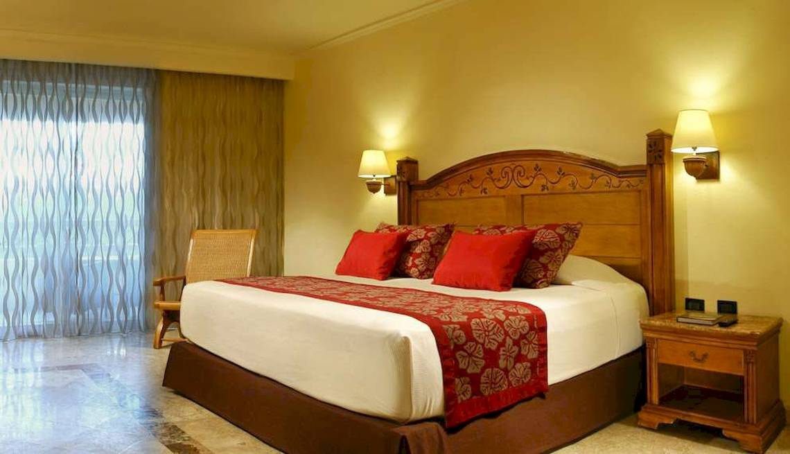 Bett im Hotelzimmer des Catalonia Royal Tulum mit rotem Kopfteil und weißen Laken.