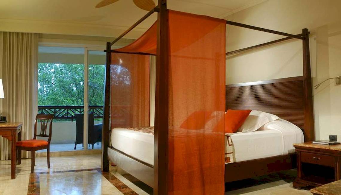 Ein Hotelzimmer mit einem Baldachin-Bett, weißem Bettlaken, orangefarbenen Kissen und einem Schreibtisch.