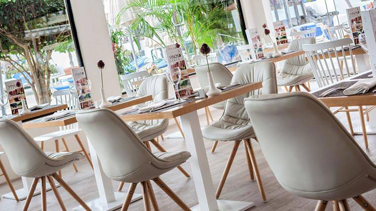 Innenansicht eines Restaurants mit weißen Stühlen und Tischen, Blick durch große Fenster auf Bäume und Autos draußen.